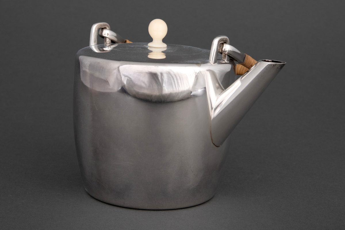 Tekanne i sølv. Sylinderformet korpus, hvorfra det reiser seg et rettvinklet håndtak med kurvfletting. På toppen er det et sirkulært lokk med lokknapp i bein, og kannens tut løper diagonalt ut fra korpus.