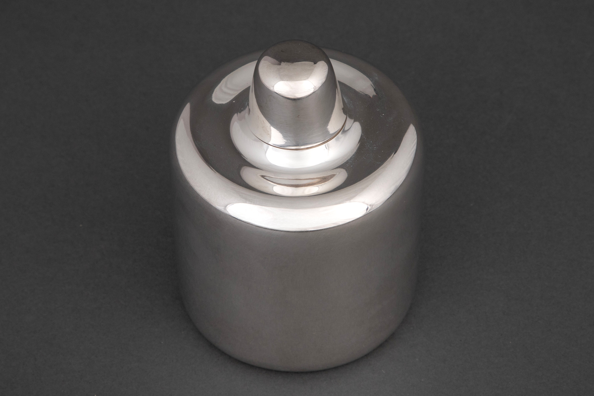 Sylinderformet tedåse i sølv med mykt avrundede kanter. Dåsen avsluttes med et høyt hvelvet lokk, som kan fungere som målebeger for en porsjon te.