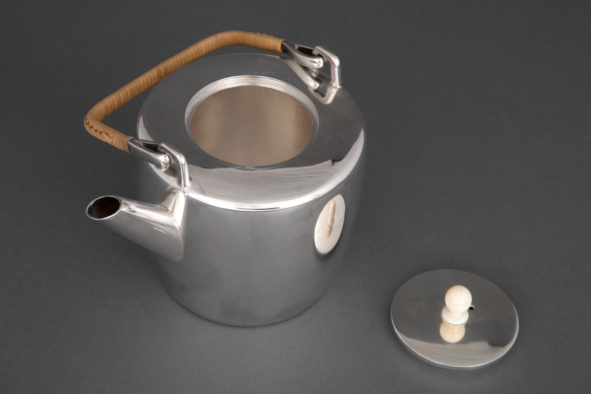 Tekanne i sølv. Sylinderformet korpus, hvorfra det reiser seg et rettvinklet håndtak med kurvfletting. På toppen er det et sirkulært lokk med lokknapp i bein, og kannens tut løper diagonalt ut fra korpus.