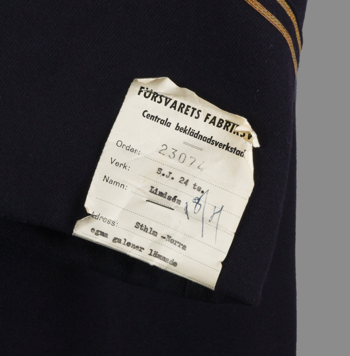 Rock av blått ylle, med sidenfoder. Dubbelknäppt med åtta knappar i gulmetall. Knapparna med krönt vinghjul tillhör 1909 års uniformsreglemente. Framtill två fickor med lock i midjehöjd. Två innerfickor i brösthöjd. Vid kragen en hank för upphängning av kavajen. Baktill sprund och slejf med två knappar. Gradbeteckning för kontorist vid ärmarna. 
Vid vänstra ärm, etikett med text: 
"FÖRSVARETS FABRIKSVERK 
Centrala beklädnadsverkstaden
Order: 23074
Verk: S.J. 24 ts.
Namn: Lindsén 18/4
Adress: Sthlm-Norra
egna galoner lämnade
Grad: Knt.
Tyg nr:
Utförande: Sidenfoder
Mått: 117-64½
Lev. tid:
Anm.:"