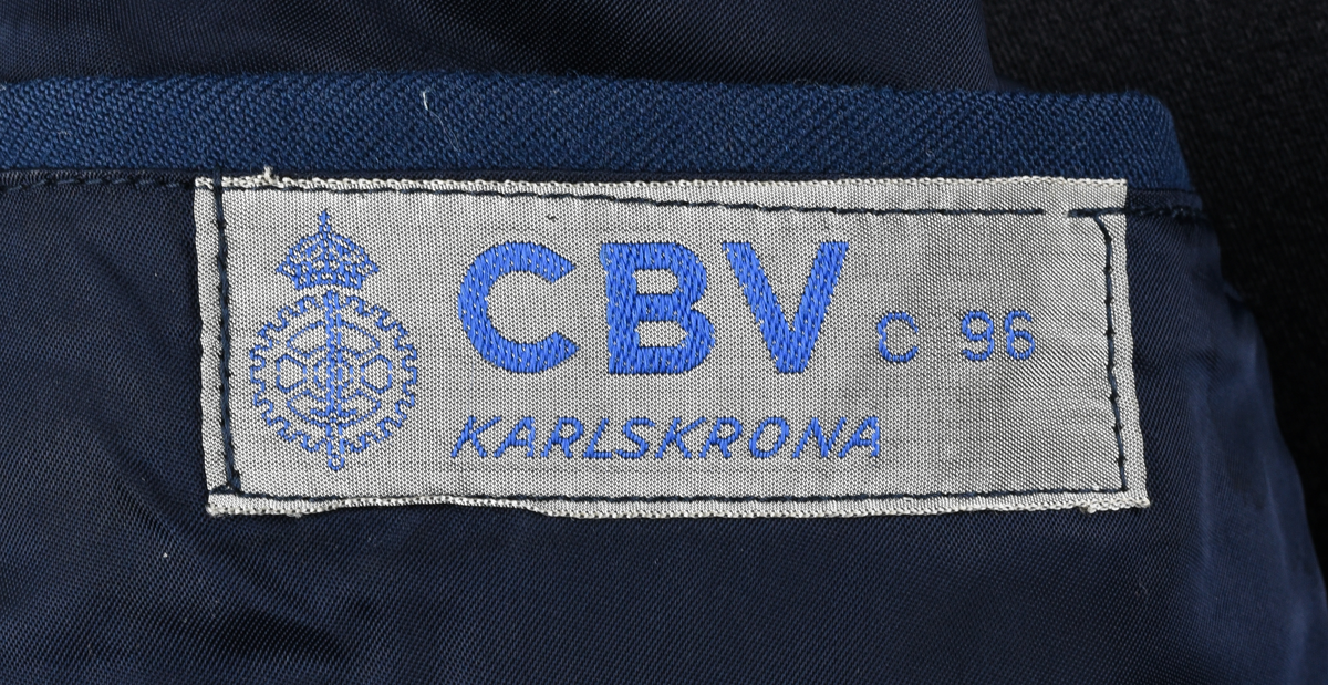 Blå kavaj, enkelknäppt med fyra knappar i gulmetall enligt 1953-års reglemente. En bröstficka utan lock och två midjefickor med lock framtill. Löstagbara axelklaffar med gradbeteckning med tre 10 mm galoner och en 7 mm galon. Vid nacken hank för upphängning av kavajen. Vid hanken fasthäftad ljusgrön lapp från kemtvätt med handskriven text "764 4/11". Två innerfickor i brösthöjd. Etikett med blå text "CBV C 96 KARLSKRONA" vid höger innerficka.