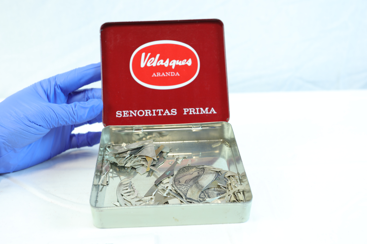 Metallskrin som er rektangulært med mørkerødt lokk. Lokket er dekorert med teksten "SEÑORITAS PRIMA" og en rød sirkel med "Velozques" og "AVANA". På fremsiden står en etikett med "20 SEÑORITAS No. 896 MADE IN HOLLAND". Skrinet ser ut til å ha vært brukt til oppbevaring av sigarer, sannsynligvis av merket Velozques. Senere har det blitt brukt av gullsmed til å samle opp sølv skroder. Lapp med "sølvs skroder" er teipet på lokket.