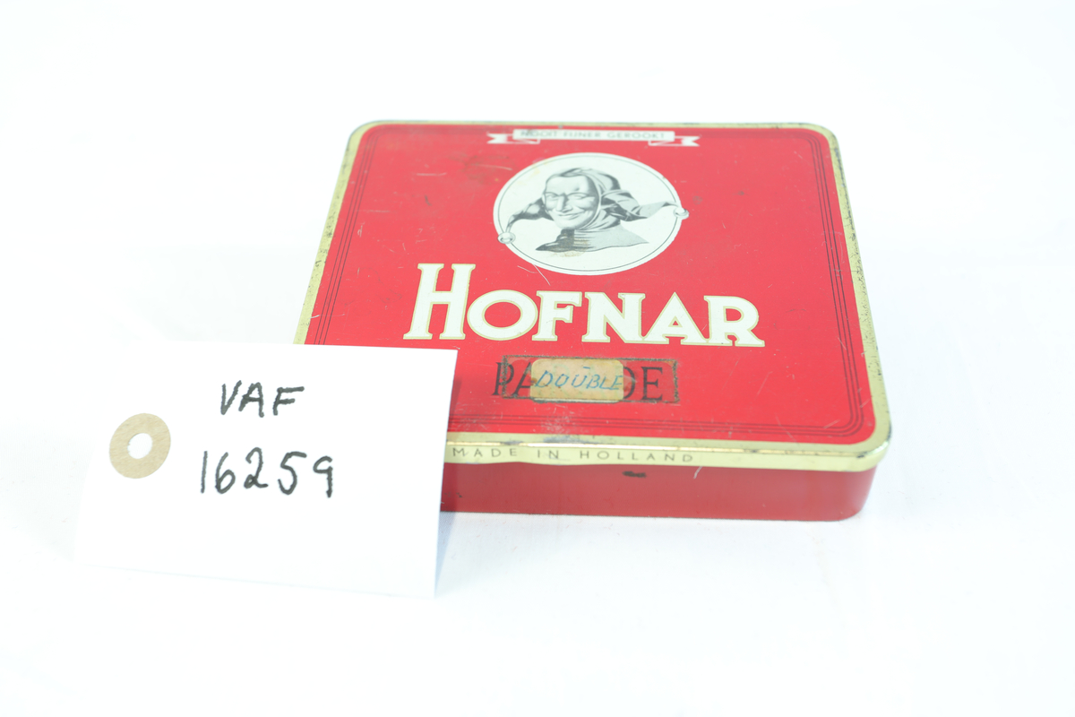 Rødt metallskrin, rektangulært med gullfargede kanter. På toppen står "HOFNAR" i store hvite bokstaver, ved siden av en illustrasjon av en mann med hatt i en hvit sirkel. Nederst "MADE IN HOLLAND". Skrinet har trolig vært brukt til tobakk eller sigarer av merket Hofnar. Nederst på skrinet står teksten "50 SIGAREN 532". Teipet på lapp hvor det står "DOUBLE". Skrinet har sannsynligvis vært brukt til oppbevaring av sigarer. Lokket på innsiden er dekorert med teksten "HOFNAR", "SIGAREN FABRIEKEN", og "PARADE", samt illustrasjoner av bygninger. I skrinet ligger det gjenstander i enten gull, kobber eller messing.