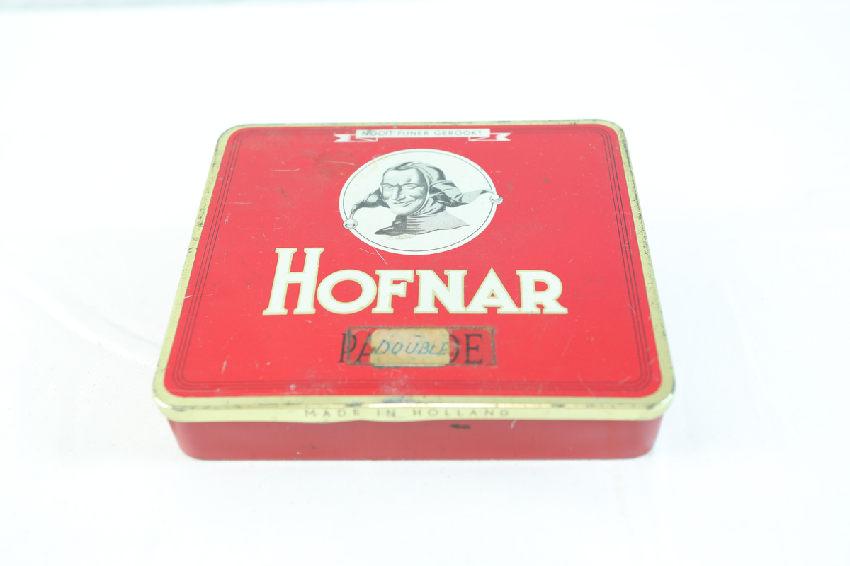 Rødt metallskrin, rektangulært med gullfargede kanter. På toppen står "HOFNAR" i store hvite bokstaver, ved siden av en illustrasjon av en mann med hatt i en hvit sirkel. Nederst "MADE IN HOLLAND". Skrinet har trolig vært brukt til tobakk eller sigarer av merket Hofnar. Nederst på skrinet står teksten "50 SIGAREN 532". Teipet på lapp hvor det står "DOUBLE". Skrinet har sannsynligvis vært brukt til oppbevaring av sigarer. Lokket på innsiden er dekorert med teksten "HOFNAR", "SIGAREN FABRIEKEN", og "PARADE", samt illustrasjoner av bygninger. I skrinet ligger det gjenstander i enten gull, kobber eller messing.