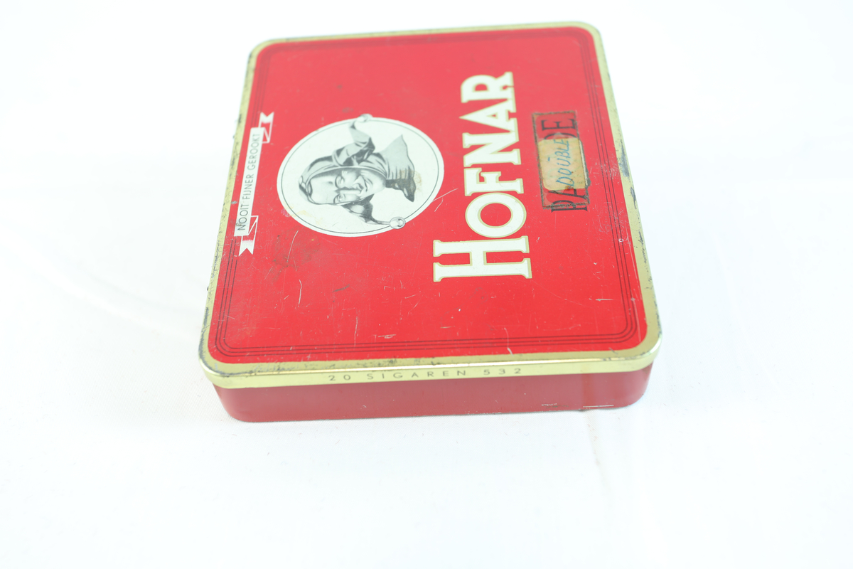 Rødt metallskrin, rektangulært med gullfargede kanter. På toppen står "HOFNAR" i store hvite bokstaver, ved siden av en illustrasjon av en mann med hatt i en hvit sirkel. Nederst "MADE IN HOLLAND". Skrinet har trolig vært brukt til tobakk eller sigarer av merket Hofnar. Nederst på skrinet står teksten "50 SIGAREN 532". Teipet på lapp hvor det står "DOUBLE". Skrinet har sannsynligvis vært brukt til oppbevaring av sigarer. Lokket på innsiden er dekorert med teksten "HOFNAR", "SIGAREN FABRIEKEN", og "PARADE", samt illustrasjoner av bygninger. I skrinet ligger det gjenstander i enten gull, kobber eller messing.