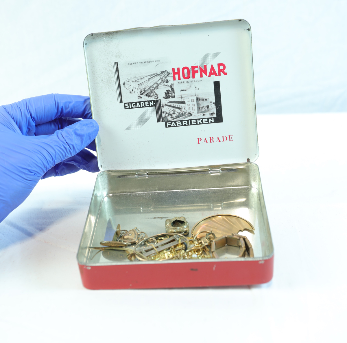Rødt metallskrin, rektangulært med gullfargede kanter. På toppen står "HOFNAR" i store hvite bokstaver, ved siden av en illustrasjon av en mann med hatt i en hvit sirkel. Nederst "MADE IN HOLLAND". Skrinet har trolig vært brukt til tobakk eller sigarer av merket Hofnar. Nederst på skrinet står teksten "50 SIGAREN 532". Teipet på lapp hvor det står "DOUBLE". Skrinet har sannsynligvis vært brukt til oppbevaring av sigarer. Lokket på innsiden er dekorert med teksten "HOFNAR", "SIGAREN FABRIEKEN", og "PARADE", samt illustrasjoner av bygninger. I skrinet ligger det gjenstander i enten gull, kobber eller messing.