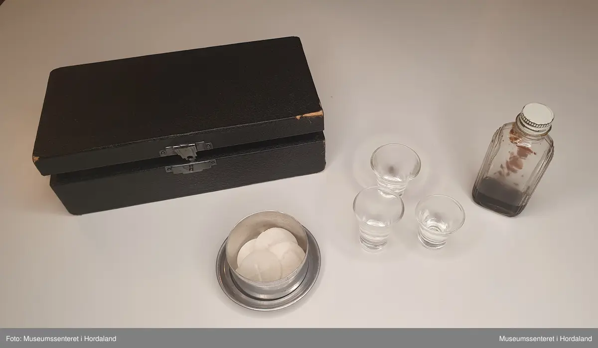 Nattverdsett bestående av et lite, svart skrin trukket med rødt for, et lite og kompakt metallfat og -skål til oblat, en liten glassflaske til vin med innhold og tre små glassbegre til altervin.
