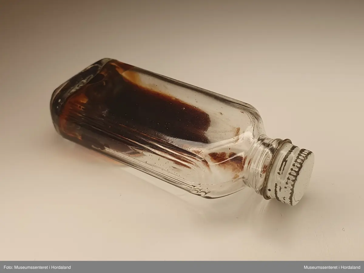 Liten, flat glassflaske med hvit skrukork av metall. Flasken inneholder mørk, stivnet væske, muligens gammel altervin.