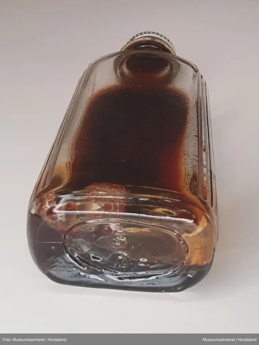 Liten, flat glassflaske med hvit skrukork av metall. Flasken inneholder mørk, stivnet væske, muligens gammel altervin.