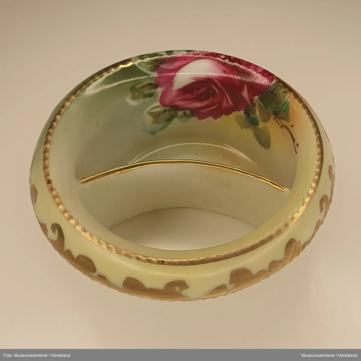 Skål av porselen med mandelformet hull på den ene siden og halvmåneformet skål på den andre siden. Den er dekorert med rose i rødt, rosa og grønt, og med forgylte detaljer.