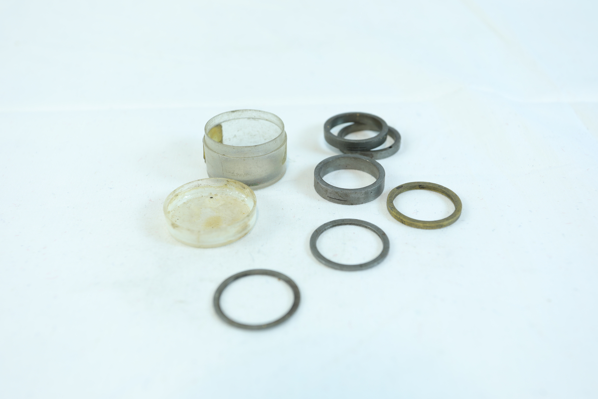 Rund liten boks i plast til oppbevaring av 7 sirkelformede metallringer. Trolig utstyr til verktøy.