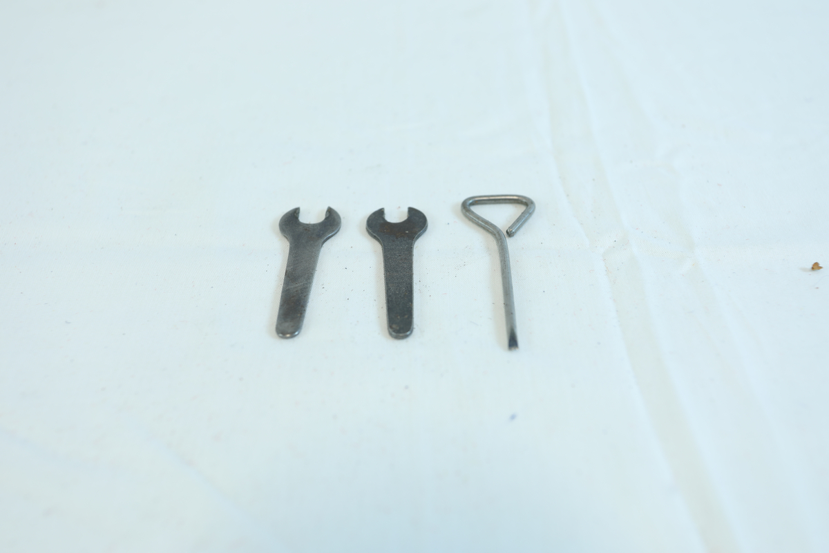 Liten plast pose, minigrip-pose, med tre redskaper i metall. Det er to fastnøkler i mørkt metall. Det siste verktøyet er sølvfarget/rustfritt stål. Den har et triangel-håndtak og tilspisset bunn. 