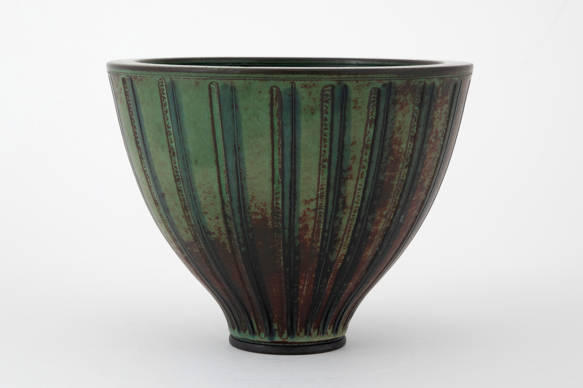 Vase i steingods. Vasen har en høy skålform med svakt utbuede sider som hviler på en markert fotring. Overflaten er dekket av brun og grønn glasur som påminner om patinert bronse. Utvendig dekorert med vertikale rifler og furer.