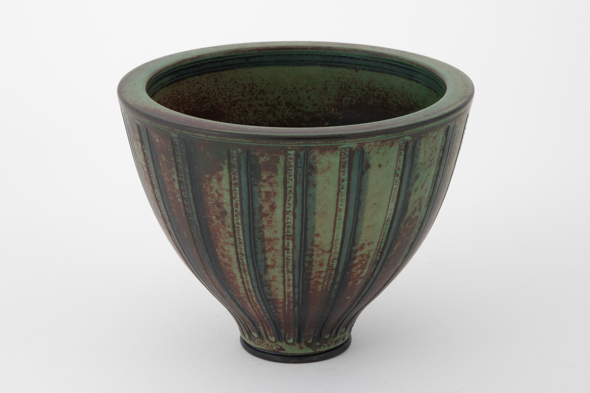 Vase i steingods. Vasen har en høy skålform med svakt utbuede sider som hviler på en markert fotring. Overflaten er dekket av brun og grønn glasur som påminner om patinert bronse. Utvendig dekorert med vertikale rifler og furer.