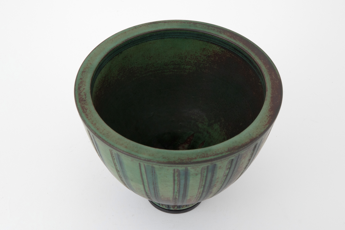 Vase i steingods. Vasen har en høy skålform med svakt utbuede sider som hviler på en markert fotring. Overflaten er dekket av brun og grønn glasur som påminner om patinert bronse. Utvendig dekorert med vertikale rifler og furer.