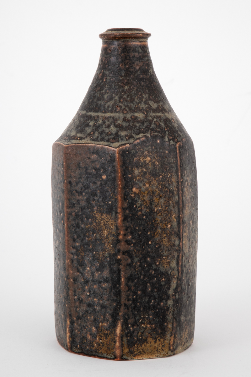 Flaskeformet vase i steingods. Vasen er formet som en oktogon, som øverst går over i en konisk hals med markert ring om den sirkelformede åpningen. Dekket av brun glasur med beigegrønne sjatteringer.
