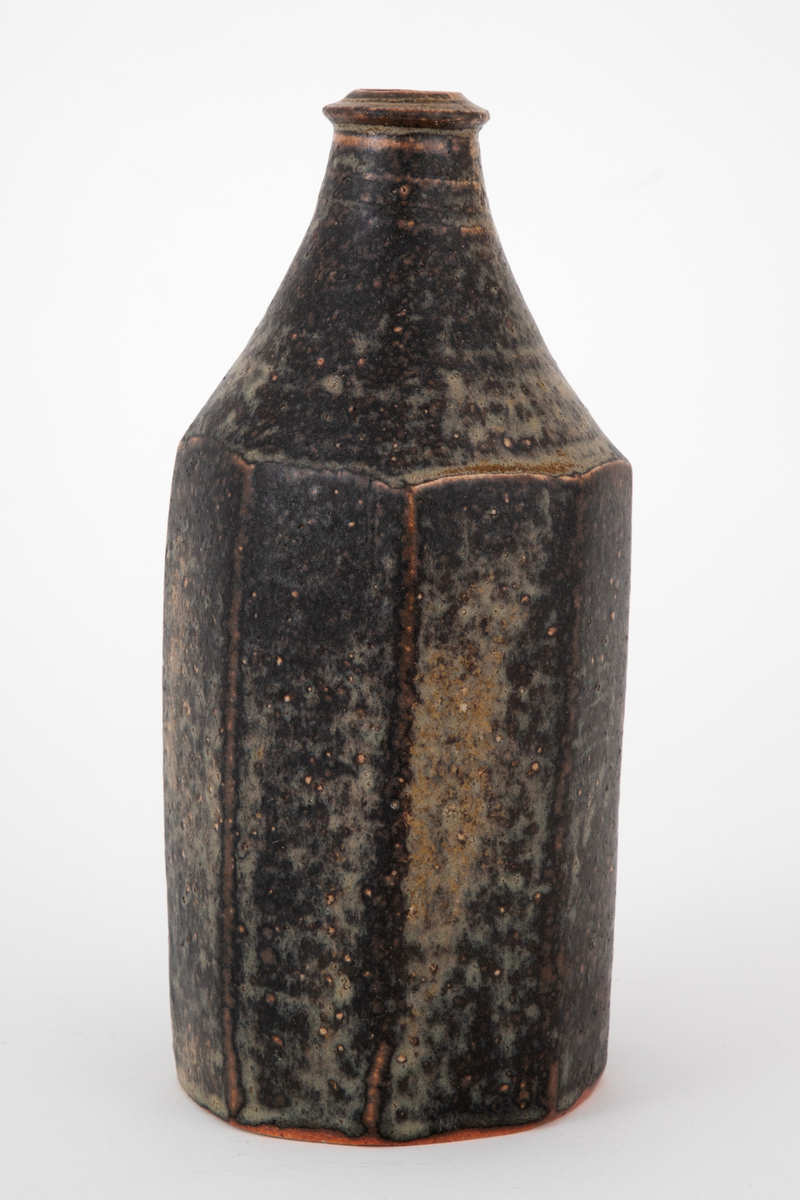 Flaskeformet vase i steingods. Vasen er formet som en oktogon, som øverst går over i en konisk hals med markert ring om den sirkelformede åpningen. Dekket av brun glasur med beigegrønne sjatteringer.