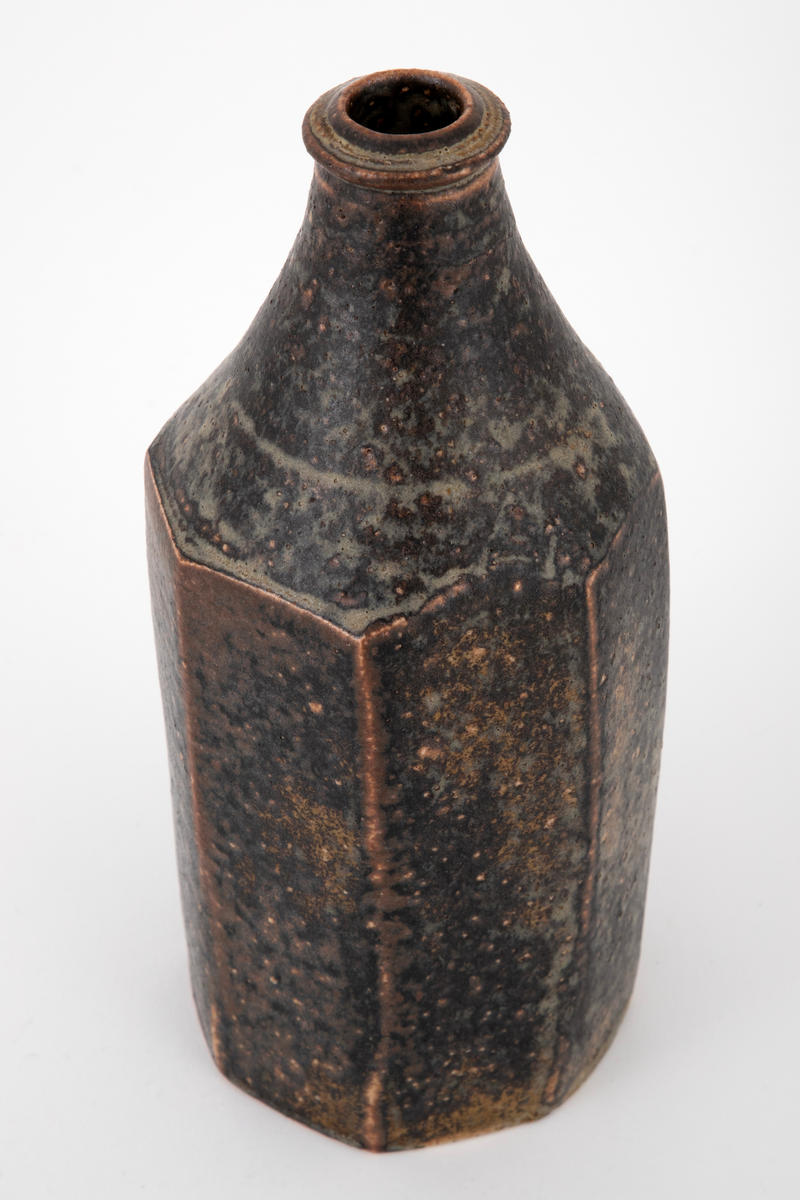 Flaskeformet vase i steingods. Vasen er formet som en oktogon, som øverst går over i en konisk hals med markert ring om den sirkelformede åpningen. Dekket av brun glasur med beigegrønne sjatteringer.