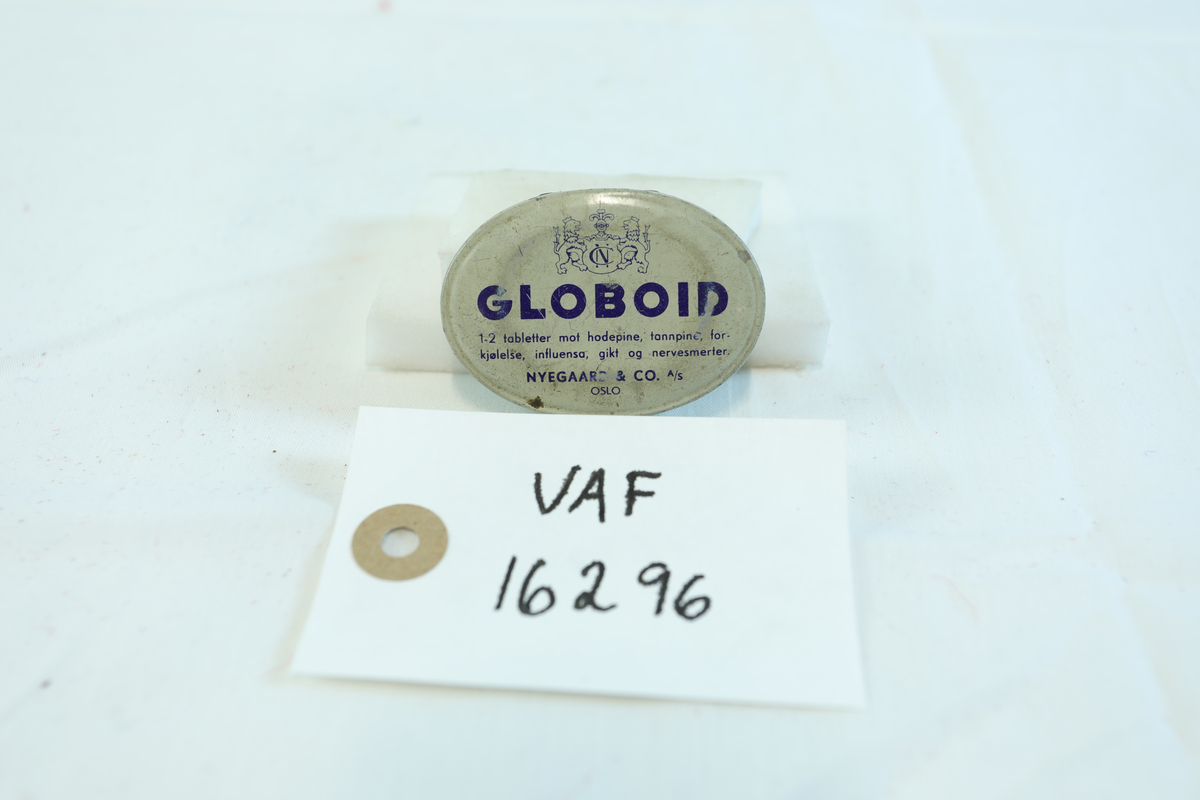 Ovalt skrin til oppbevaring av globoid fra Nyegaard & Co A/S. Skrift på både forside og bakside. Skrinet inneholder en flat klump med metall. 