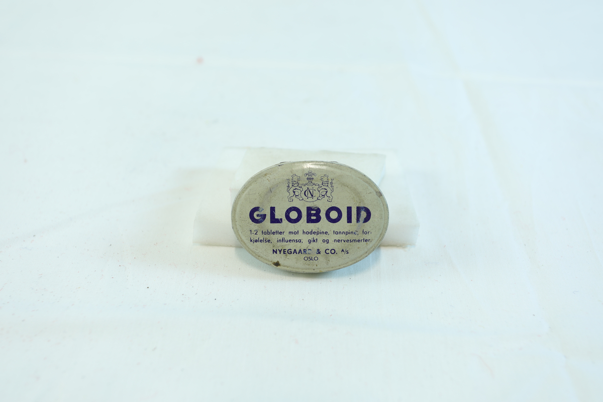 Ovalt skrin til oppbevaring av globoid fra Nyegaard & Co A/S. Skrift på både forside og bakside. Skrinet inneholder en flat klump med metall. 