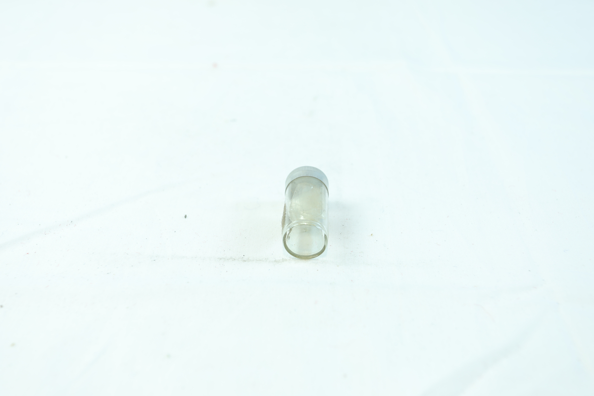 Lite glass med lokk i metall. I beholderen er det sammenbrettet gjennomsiktig plass med en liten perleformet gjenstand. 