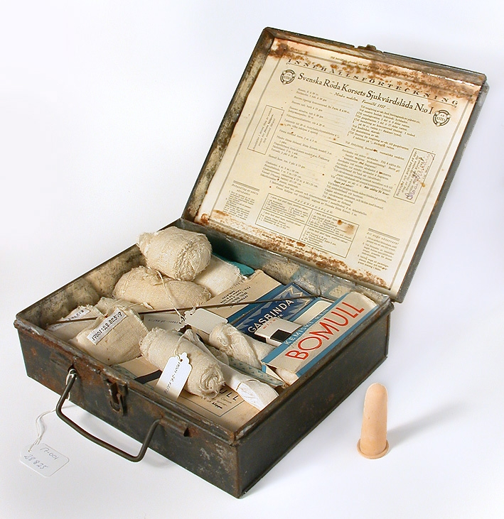 Låda av svart plåt med handtag. Papper med text på insidan locket och i botten. Från Bomstastugan byggd 1928.

2-7 Gasbindsrullar, 8-9 Tygremsrullar, 10 Förpackning med bomull, Astra, 11 
Förpackning med bomull. Text: "Patrull Björen", 12 Bunt med vita band, 13 Förpackning gasbinda Wilhelm Bäckman, 14 Förpackning gasbinda Blå kartong, 15-16 Förpackning gasbinda Mölnlycke, 17 Förpackning första förband, 18 Fingerskydd ur gummi.