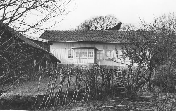 Vrå gård i Vallda. Bostadshus med glasveranda och ekonomibyggnad till vänster. Vid trådstaketet i förgrunden pågår häckklippning via stege. När fotografiet togs ägdes gården av Arvid och Josefine Källberg. De hade sonen Arnold (f. 1927). Gården var Josefines hem och kvar bodde även Josefines syster Bernhardina Andersson och fosterbror Torsten Andersson. Föräldrarna hette König och Augustina Andersson (f. Pettersson), vilka i sin tur övertagit den efter hennes föräldrar Magnus (f. 1818) och Maja Johanna Pettersson (f. Carlsson 1816). Gården var i släktens ägo fram till 2013. 
(Se även bild G8369).