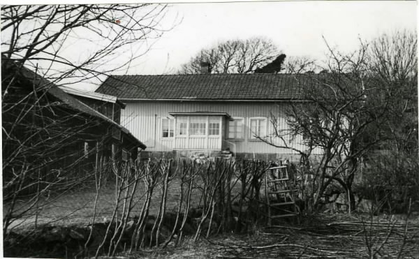 Vrå gård i Vallda. Bostadshus med glasveranda och ekonomibyggnad till vänster. Vid trådstaketet i förgrunden pågår häckklippning via stege. När fotografiet togs ägdes gården av Arvid och Josefine Källberg. De hade sonen Arnold (f. 1927). Gården var Josefines hem och kvar bodde även Josefines syster Bernhardina Andersson och fosterbror Torsten Andersson. Föräldrarna hette König och Augustina Andersson (f. Pettersson), vilka i sin tur övertagit den efter hennes föräldrar Magnus (f. 1818) och Maja Johanna Pettersson (f. Carlsson 1816). Gården var i släktens ägo fram till 2013. 
(Se även bild G8369).