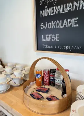 Benkeplate med kaffekopper, asjetter og en kurv med Vestlandslefser, brusbokser og -flasker. En meny-tavle på veggen over.over 