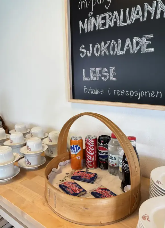 Benkeplate med kaffekopper, asjetter og en kurv med Vestlandslefser, brusbokser og -flasker. En meny-tavle på veggen over.over 