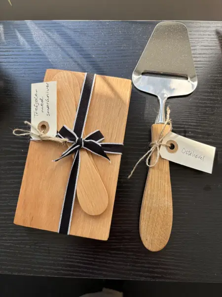 En ostehøvel med treskaft ved siden av en trefjøl med to smørkniver oppå ligger på en sort hylle.