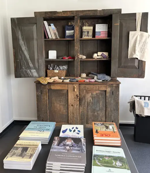 Et bord med seks bokstabler på i forgrunn. I bakgrunn et gammelt framskap med bøker og småvarer i hyllene.