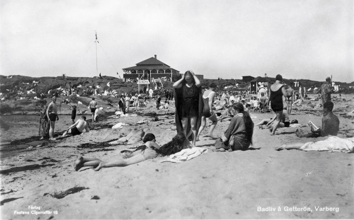 Vykort, "Badliv å Getterön, Varberg". Myllrande strandliv år 1926 i 4:e vik där en kiosk står till höger och uppe på berget ligger Sundhs restaurang. Till vänster står en man och ler mot den okände fotografen.