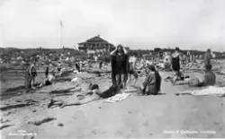Vykort, "Badliv å Getterön, Varberg". Myllrande strandliv år 1926 i 4:e vik där en kiosk står till höger och uppe på berget ligger Sundhs restaurang. Till vänster står en man och ler mot den okände fotografen.