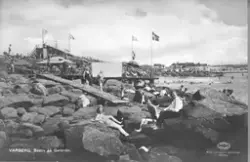 Vykort, äkta fotografi, "Varberg. Badliv på Getterön." Badliv i 4:e vik, sett mot land med solbadare vid Ståhlbrands brygga med café och rutschkana. I bakgrunden ses byggnader inne i viken. 1930-talet.