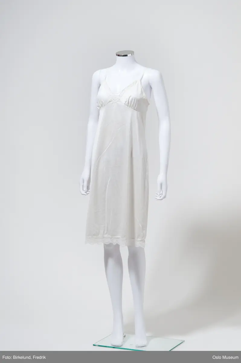 Hvit underkjole i blank tekstil med stropper, blondekant i hals og nederst. Fra 1950-tallet.