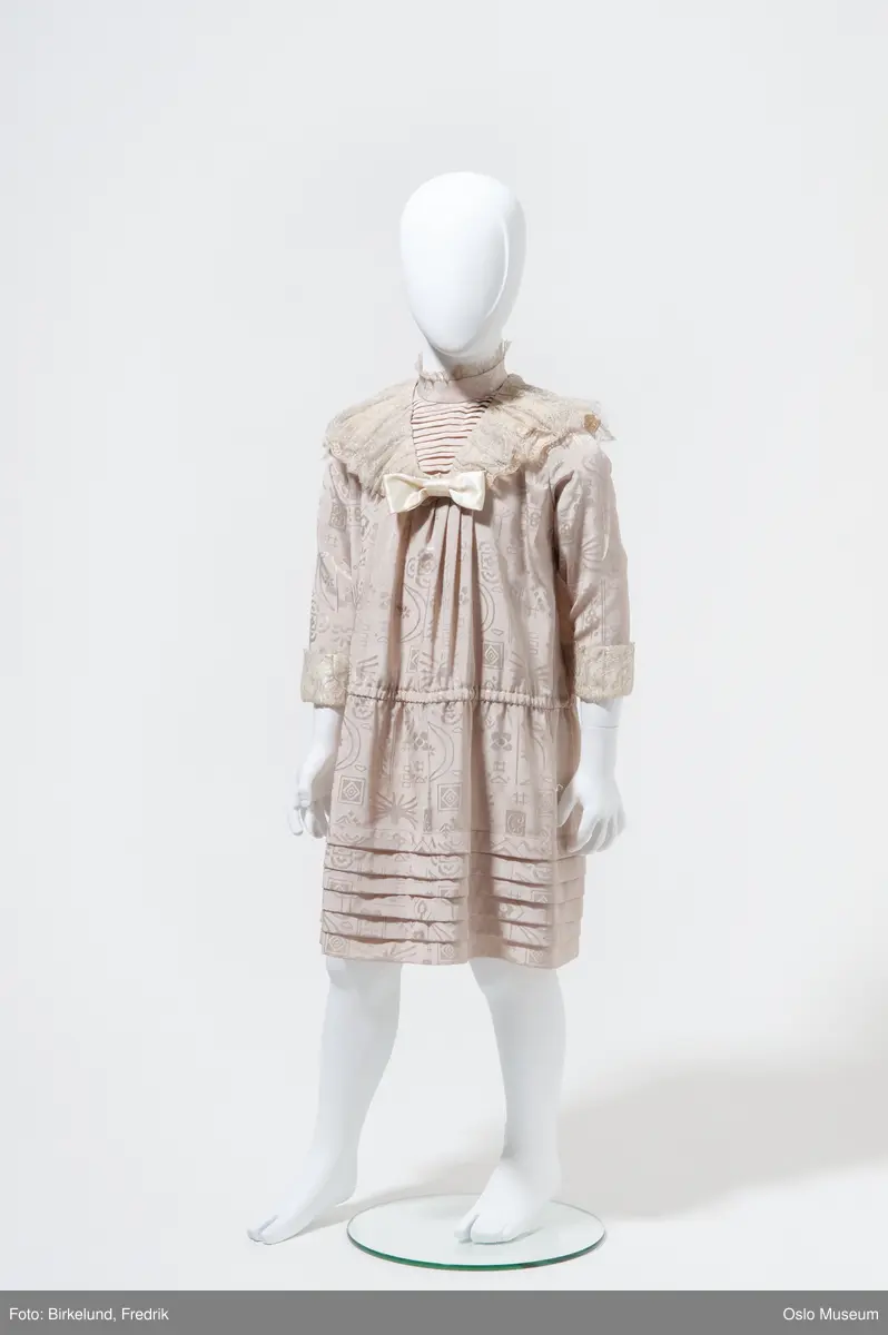 To identiske kjoler i barnestørrelse i eggeskallhvitt brokadeliknende stoff med hvit avtagbar blondekrave og silkesløyfe i halsen. |1||