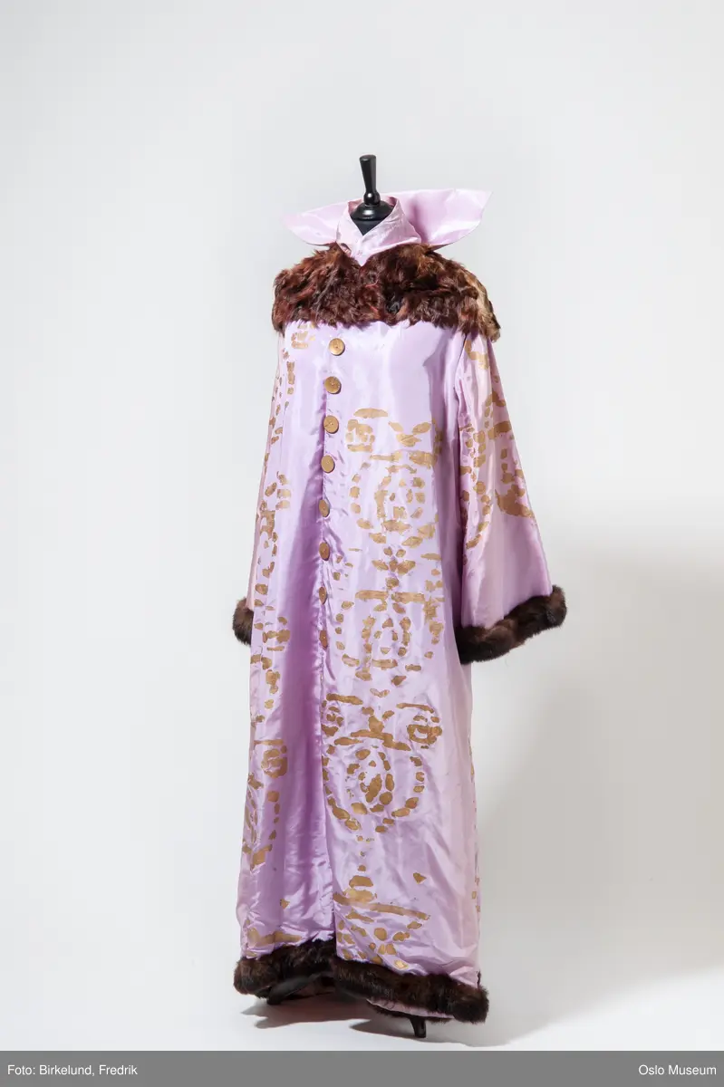 Kåpe i lys rosa silkestoff med brun skinnkrage, skinnkant på ermer og nederste kant. Dekor i form av påmalt gullmønster. Lukning i form trykk-knapper innvendig, utvendig i form av store gullmalte knapper. For av hvitt stoff.
