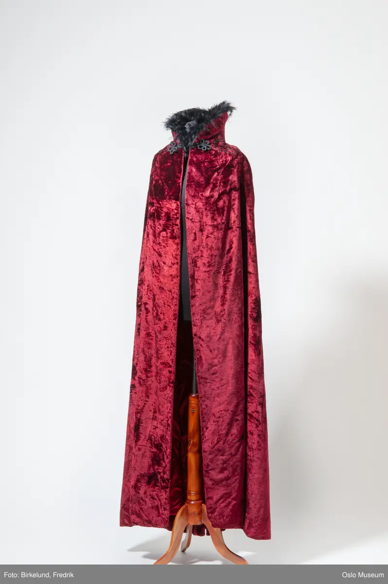 Lang cape i mørkerødt fløyelsstoff med mørk brun krage av fuskepels, lukning i form sort hempe øverst. For av rød silke