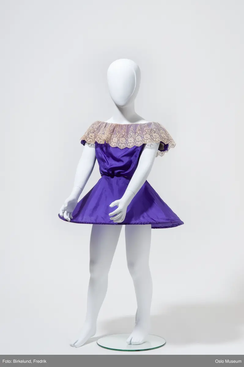 Lilla ballettkostyme, kjole, i silke (?), kortermet, med blondekrage. I skjørte-del er innsydd en metall sirkel slik at skjørtet står ut av seg selv. 