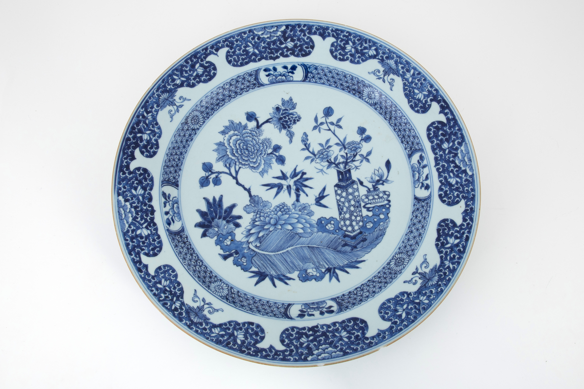 Rundt fat i keramikk. Dekket med hvit glasur med blålig skjær. Dekorert med malt, blå underglasur. I speilet ses et innrammet blomstermotiv. Rundt kanten en bord med blant annet blomster. Smal lysebrun kant.