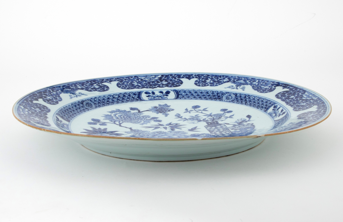 Rundt fat i keramikk. Dekket med hvit glasur med blålig skjær. Dekorert med malt, blå underglasur. I speilet ses et innrammet blomstermotiv. Rundt kanten en bord med blant annet blomster. Smal lysebrun kant.