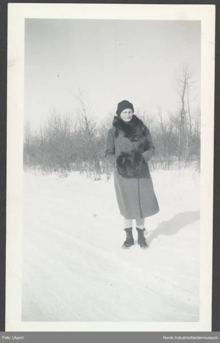 Gunhild Overland kledd i vinterkåpe med hendene i pelsmuffe og pelskrage av rev rundt halsen. Stående i snødekket landskap.