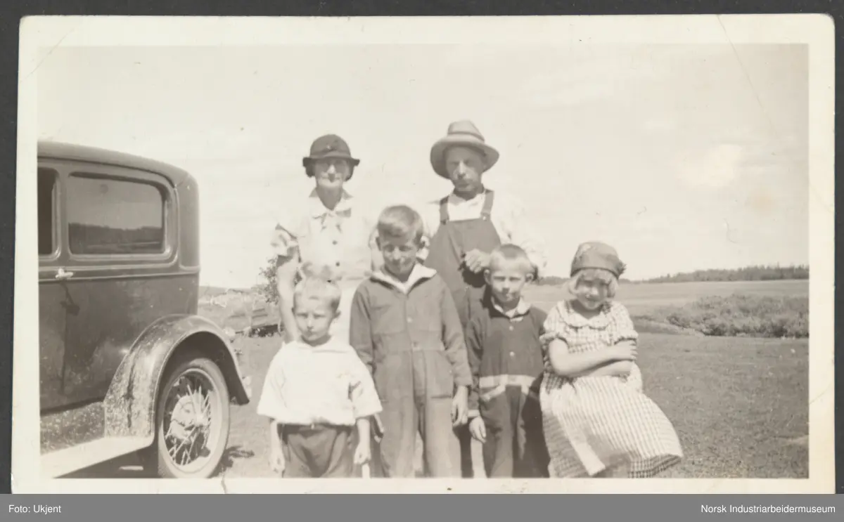 Familien Overland stående landbrukslandskap i Preeceville. Familie med fire barn. Bil stående på siden.