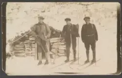 Tre menn på skitur. En av mennene med solbriller, alle har sekk på ryggen. Laftet løe i bakgrunnen.