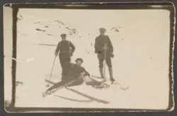 Tre menn på skitur i fjellandskap. En av mennene ligger i snøen med ski på beina.