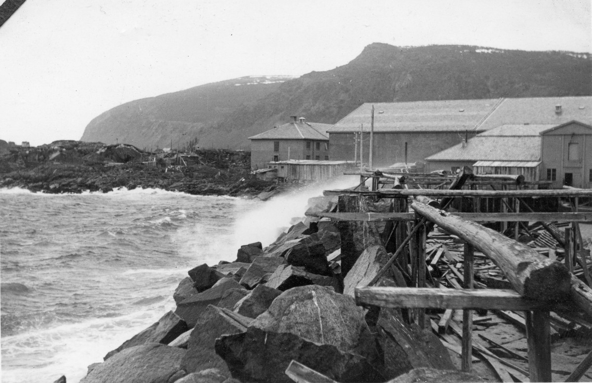 Dårlig vær og sjøen pisker oppover moloen nedenfor Filetfabrikken. «Sosialen» ses midt i bildet. 

Haugnyken og Vardheia (Haugheia) i bakgrunnen.   
