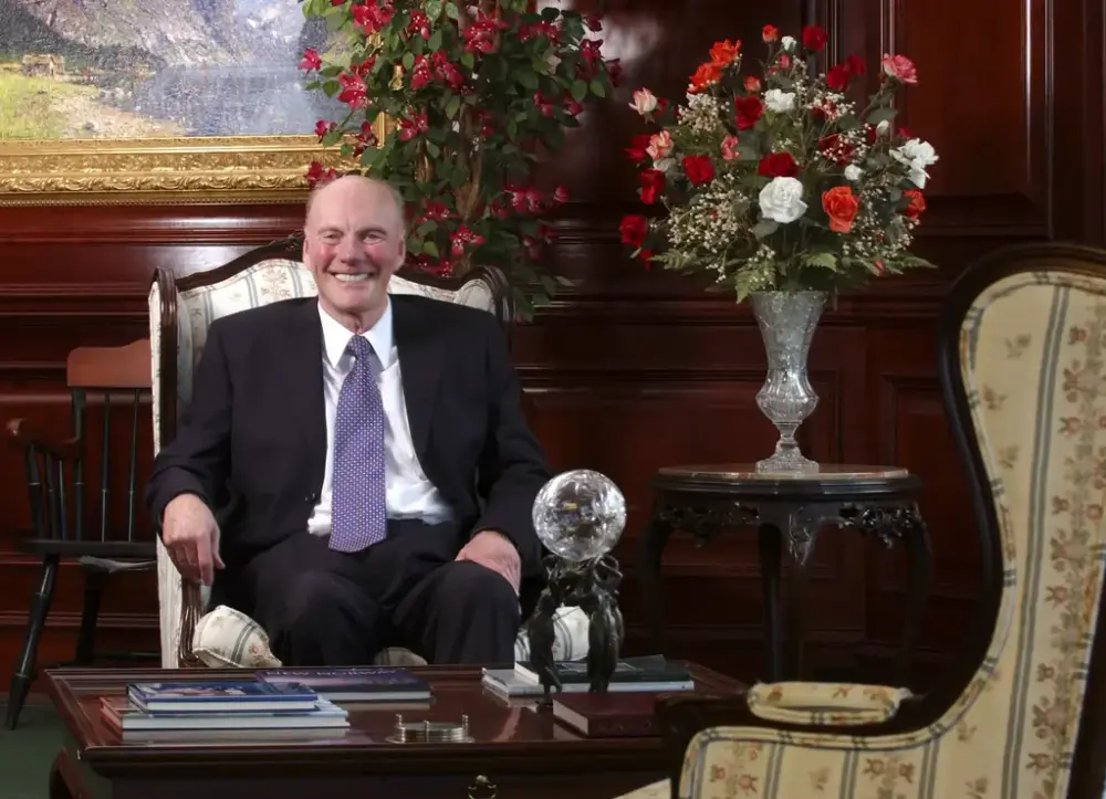 En mann med stort smil og slips i et overklasseinteriør med store blomsteroppsatser i bakgrunn.