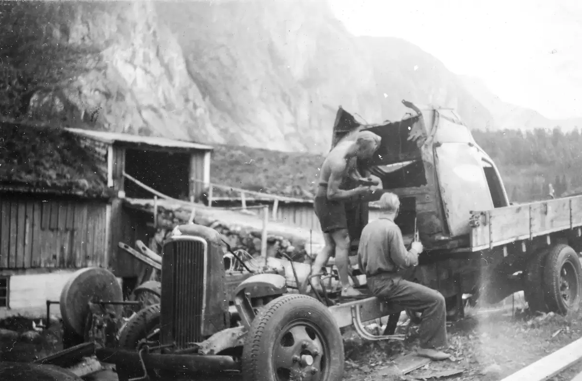 Historisk bilde av to menn som reparerer en lastebil. Fjell i bakgrunn.