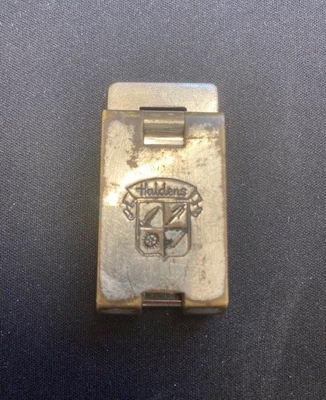 Sammenleggbar lupe i etui av lær. Lupen foldes opp slik at områder på 5mm og 10mm og en tomme vises i synsfeltet. En kan så telle hvor mange tråder er på dette området, for å kvalitetssikre tekstilet. På stativets utside er logo fra Haldens gravert i overflaten.